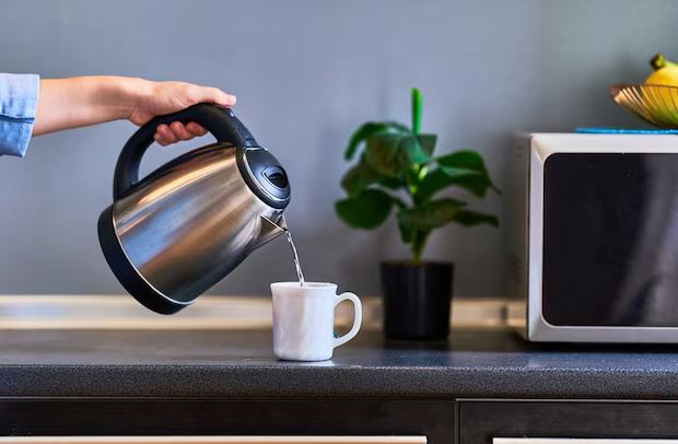 Chaleira elétrica inox sobre a bancada de cozinha, com vapor saindo do bico e xícaras de chá ao lado.
