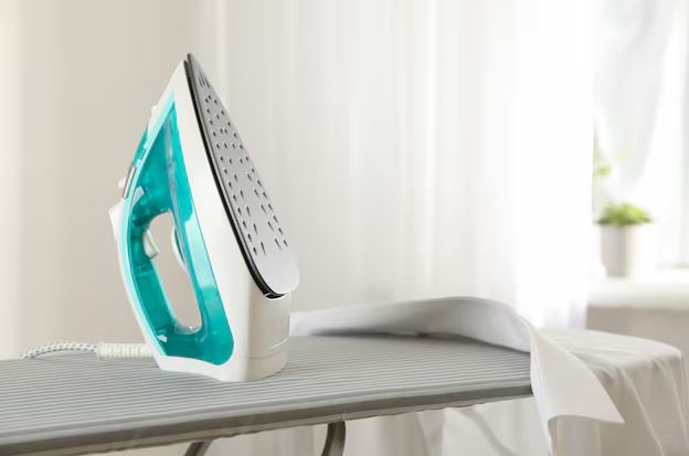 Ferro de passar roupa em uso sobre uma tábua, em área de lavanderia residencial, com peças de roupa ao lado.