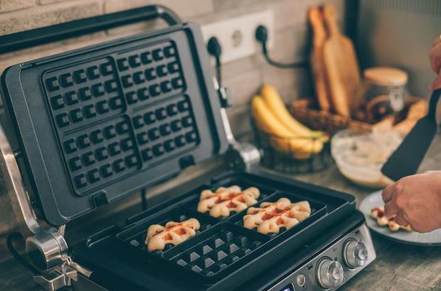 Máquina de waffle aberta sobre a bancada com waffles dourados recém-preparados e acompanhamentos ao redor.