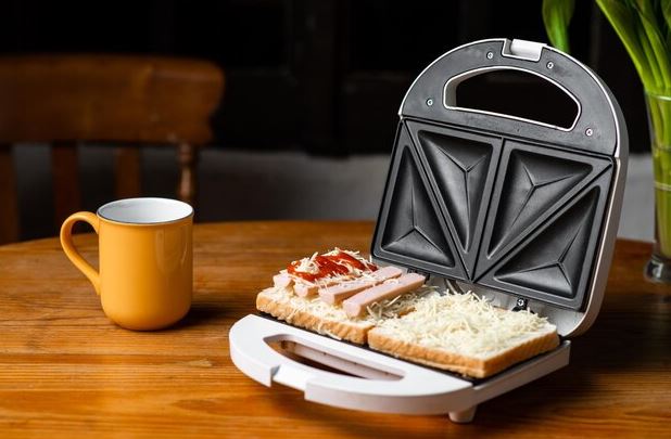 Sanduicheira grill aberta em bancada de cozinha, com sanduíches dourados sendo preparados ao lado de ingredientes frescos.