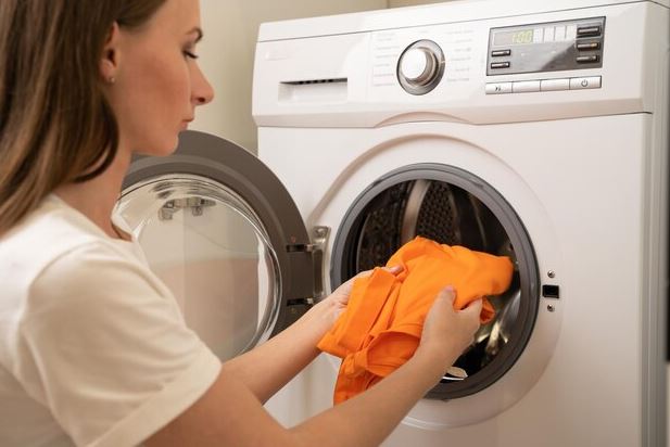 mãos colocando roupas úmidas em uma secadora em uma lavanderia compacta de apartamento, ao lado de um cesto de roupas