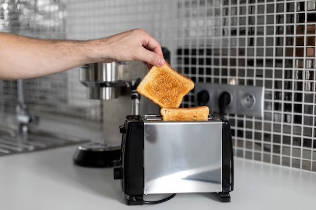 Torradeira moderna em bancada de cozinha, com fatias de pão douradas saindo das fendas ao lado de um café da manhã servido.
